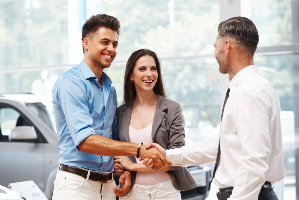 Car Loans Banner - Young couple shaking hands with sales representative at a dealership after buying a new car