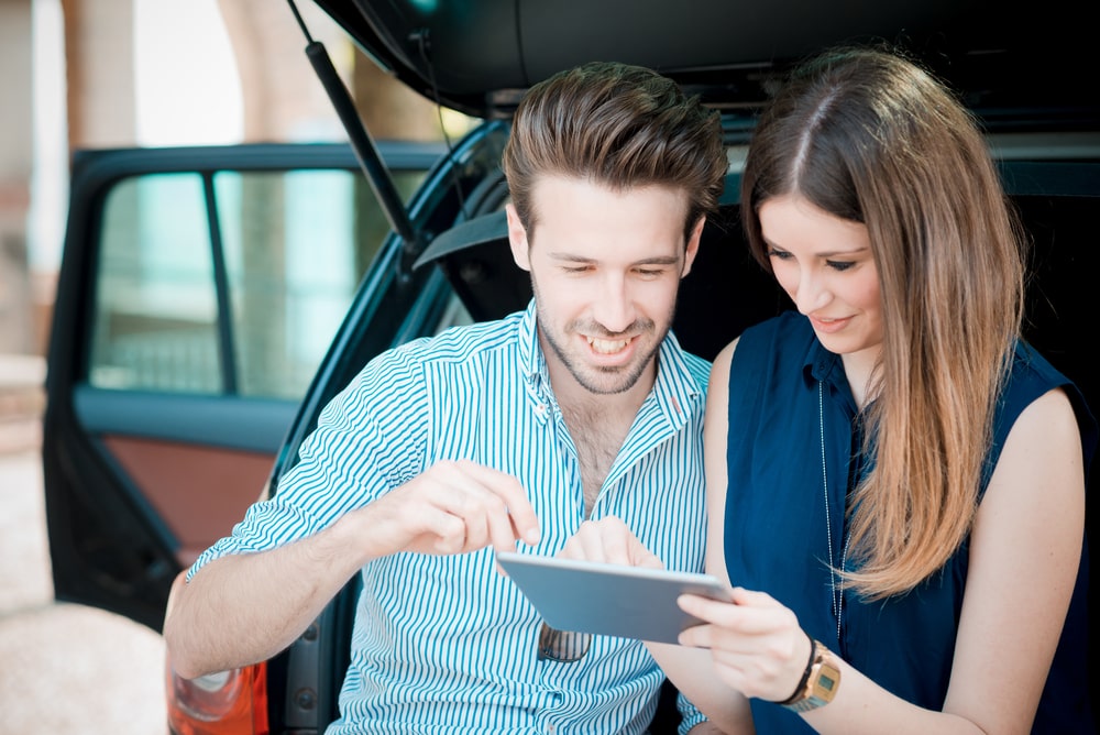 Car Loans Banner - Young couple looking at a tablet while sitting in the boot of their car