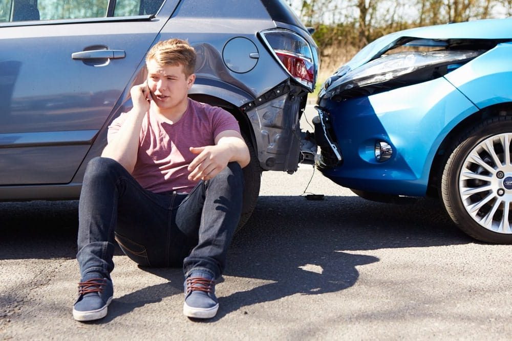 Car Insurance Banner - Man sitting next to a car accident talking on the phone to an insurance company to make a claim.
