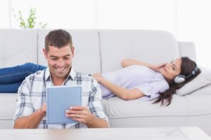 Health Insurance Banner - Man smiling looking at his tablet while a woman lies on the couch with headphones on