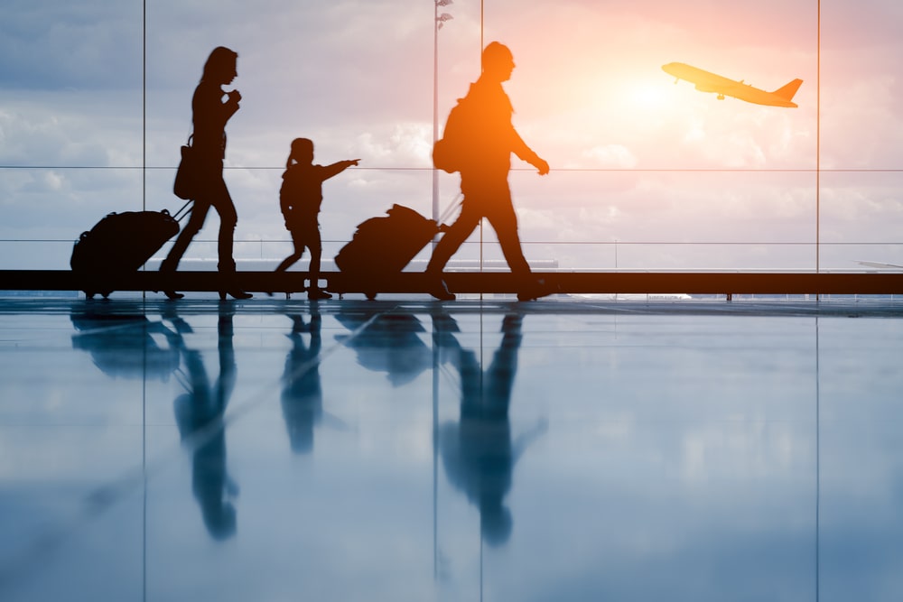Travel Insurance Banner - Wide shot of a family in silhouette walking through an airport next to the window.