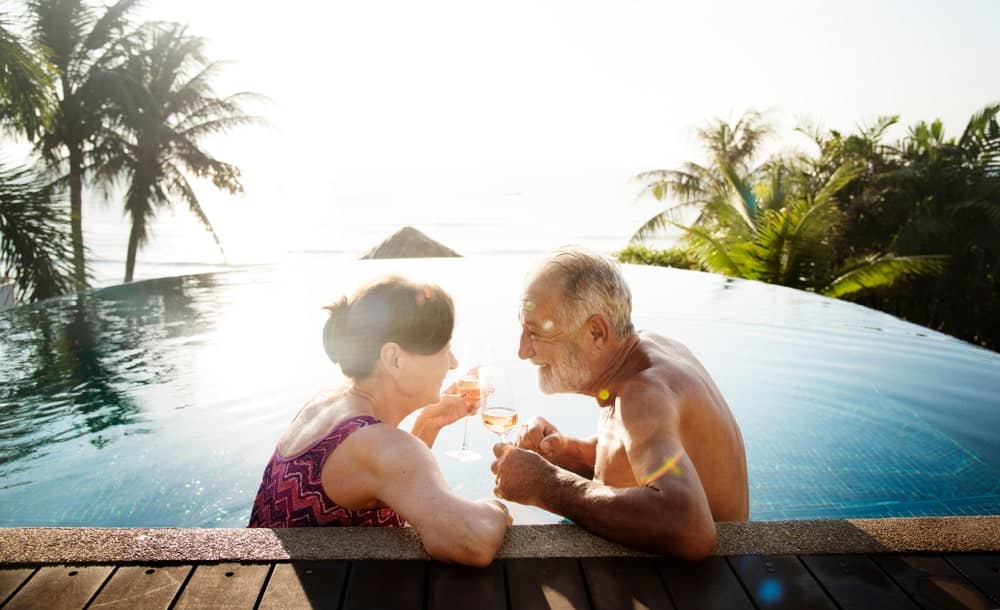 Travel Insurance Banner - Senior couple drinking wine in a pool overlooking a tropical landscape.
