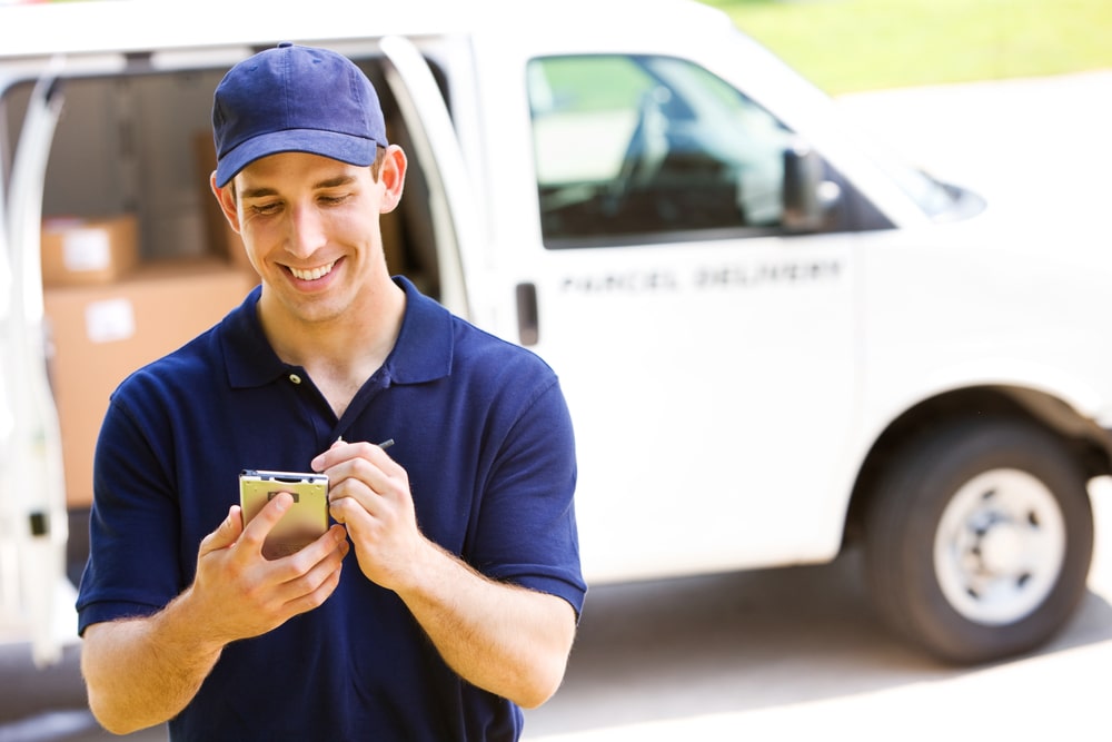 Car Insurance Banner - A delivery driver standing in front of his van checking an order off on his device.