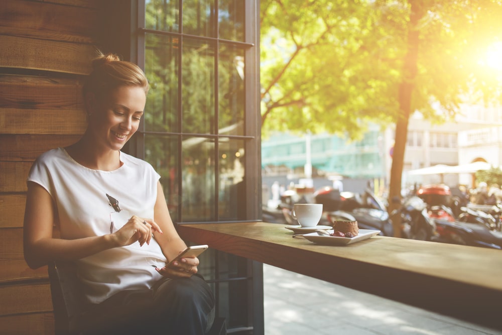 Life Insurance Banner - Smiling woman looking at her phone while sitting at the window of a cafe