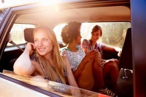 Car Insurance Banner - Young woman smiling while looking out the window of a car next to a group of friends