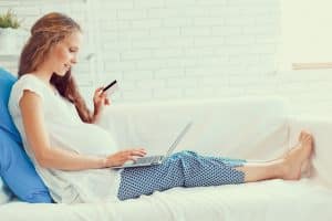 Money Transfer Banner - Pregnant woman lying on the couch looking at her laptop while holding a credit card