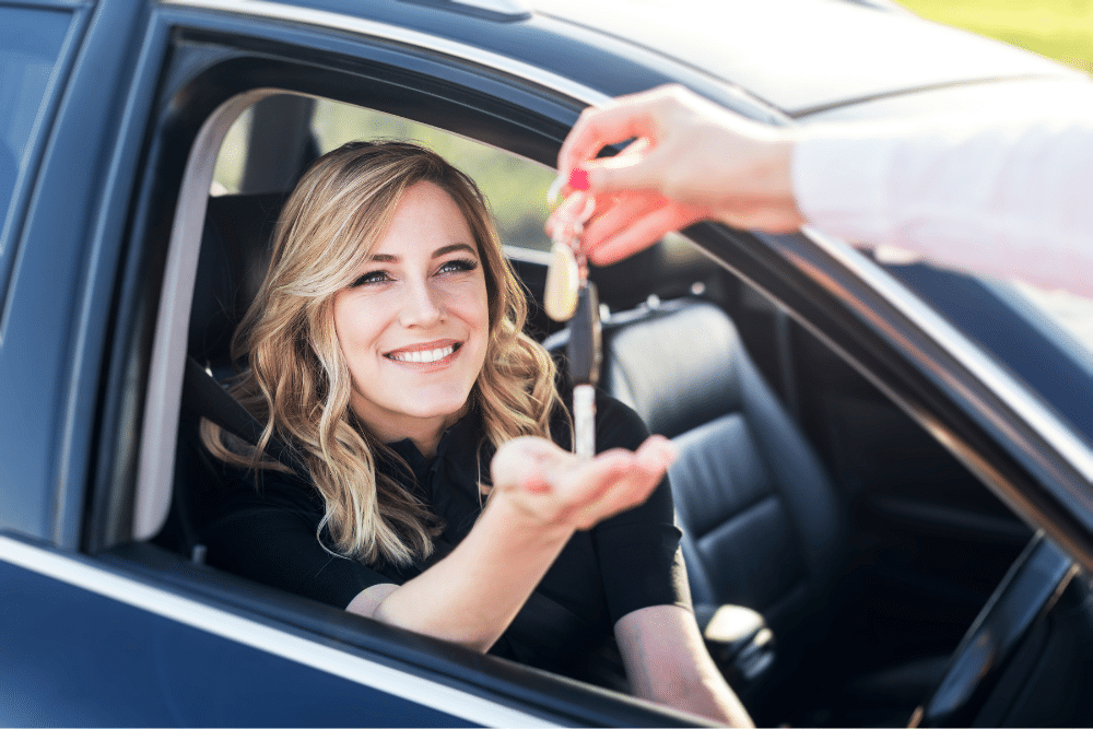 Car Loans Banner - Woman sitting at the wheel of her new car, receiving the keys from the seller