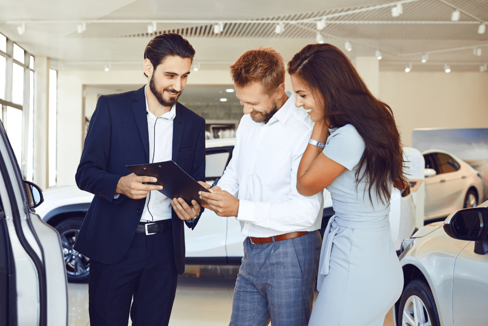 Car Loans Banner - Couple signing a contract to buy a car at a dealership with a salesperson